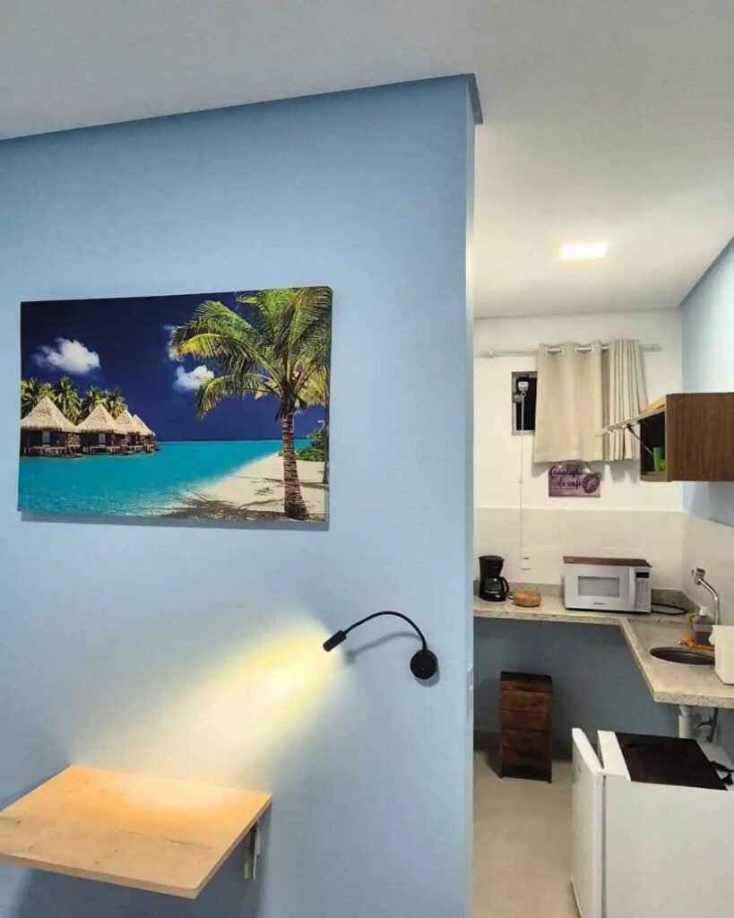 Bancada de trabalho e leitura com luminária articulada acesa e quadro praiano na parede azul.