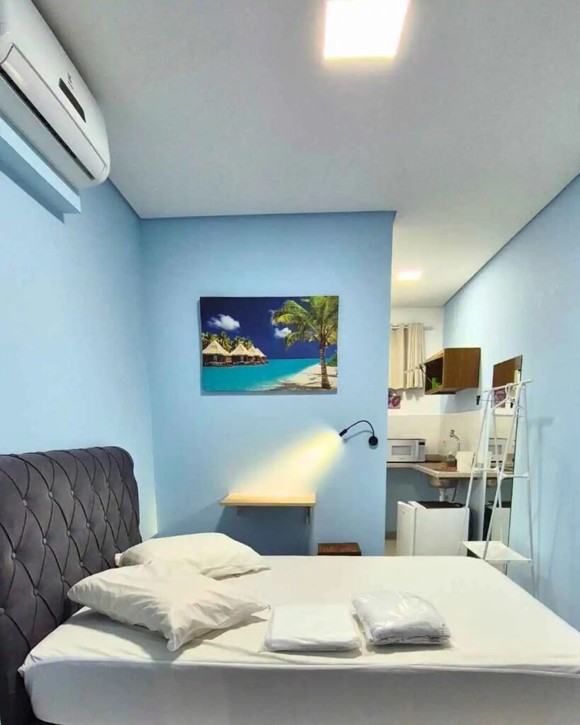 Studio acomodação com cama de casal estofada, enxoval limpo e ambiente integrado com mini copa ao fundo.
