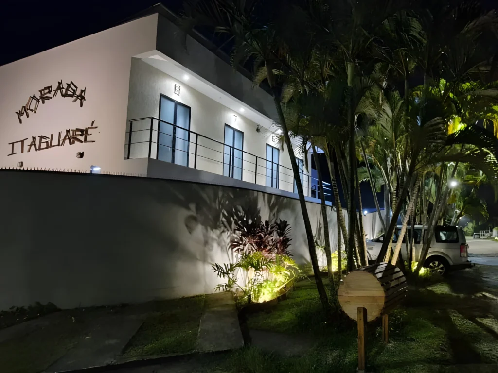 Entrada iluminada da Morada Itaguaré à noite com destaque para as palmeiras e arquitetura.